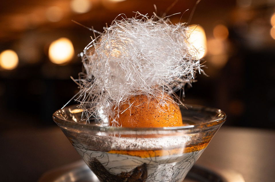 A golden sea buckthorn sphere topped with a delicate ball of spun sugar, served in an etched martini glass, against a warm bokeh background.