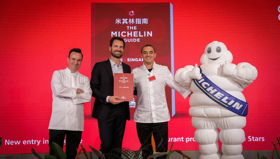 Gwendal Poullennec in a dark blazer holds a red Michelin Guide Singapore book, flanked by two chefs in white jackets, standing in front of a large red Michelin Guide Singapore backdrop and an inflatable Bibendum mascot at a star announcement ceremony.