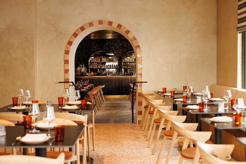A warmly lit dining room with arched brick-trimmed doorways, light wood chairs, dark tabletops set with glassware and amber tumblers, and a bar