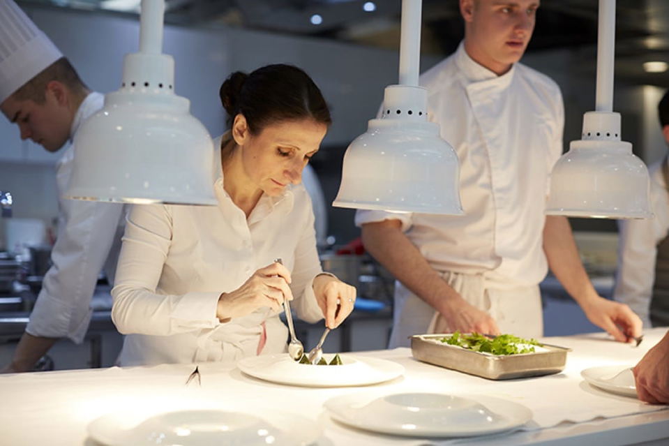 Anne-Sophie Pic, a chef in a white uniform, meticulously plates a dish with tongs and a spoon under bright heat lamps in a professional kitchen.