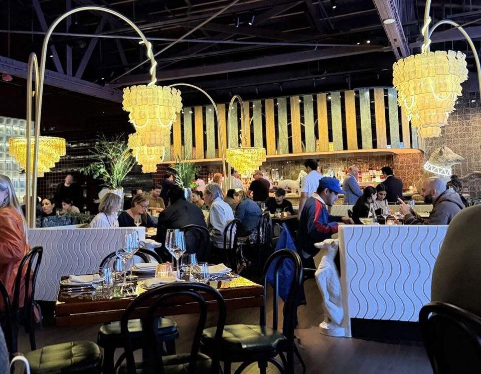 A busy, upscale restaurant dining room with ornate gold chandelier-style pendant lights, white wave-textured booth dividers, and a bar in the