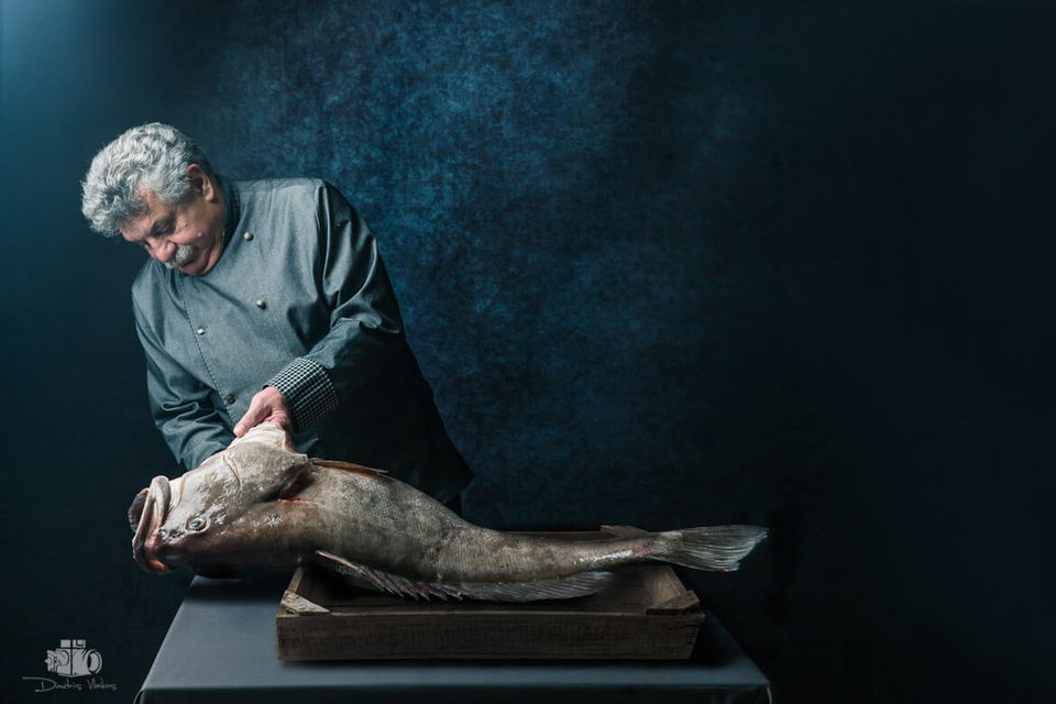 Lefteris Lazarou, a chef with grey hair and a mustache, in a grey chef's jacket, holds a large whole fish over a wooden crate.