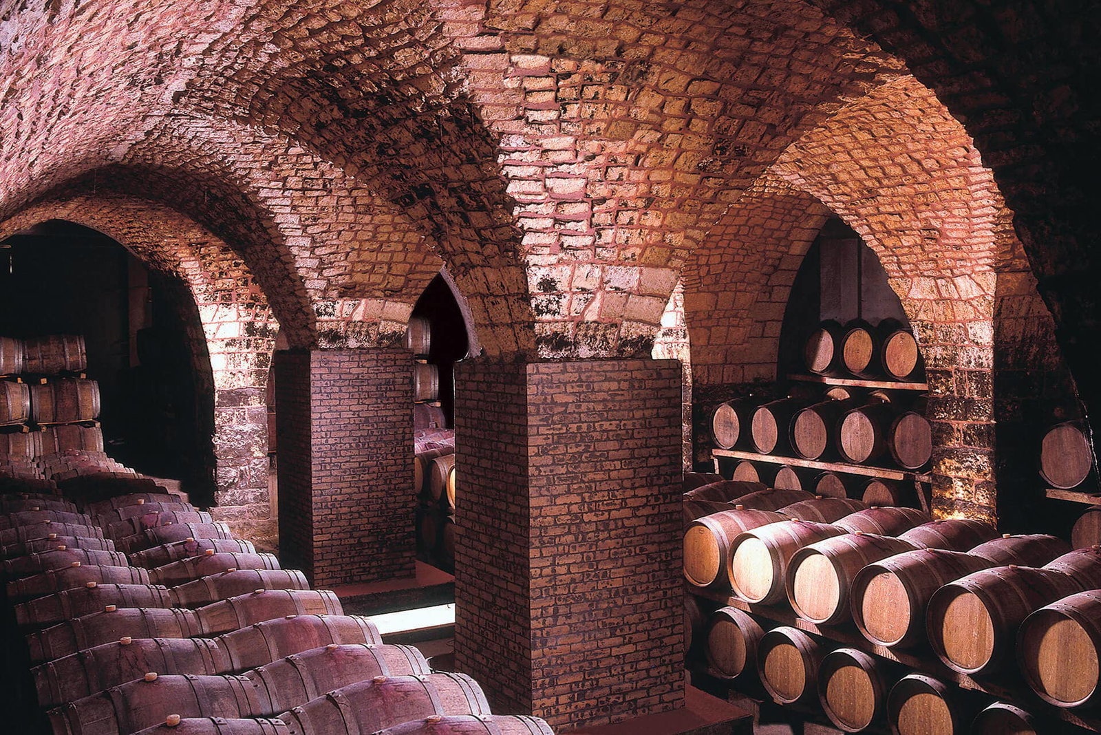A historic stone-vaulted wine cellar with rows of oak aging barrels stacked on wooden racks, creating a deep perspective.