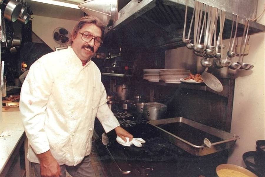 Chef Alain Borel, wearing a white chef coat and glasses, wipes down a stovetop in the kitchen at L'Auberge Provençale, with hanging ladles and stacked plates visible behind him.