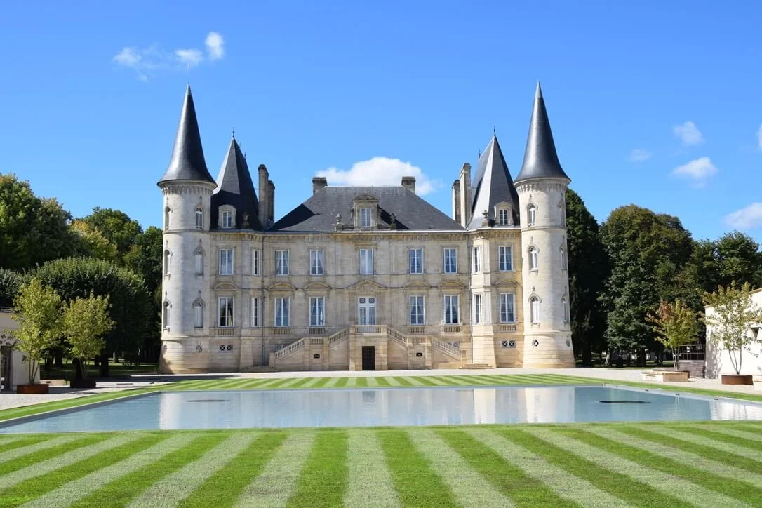 The grand exterior of Château Pichon Baron, a four-towered limestone château with conical slate roofs, a formal striped lawn, and a reflecting pool.