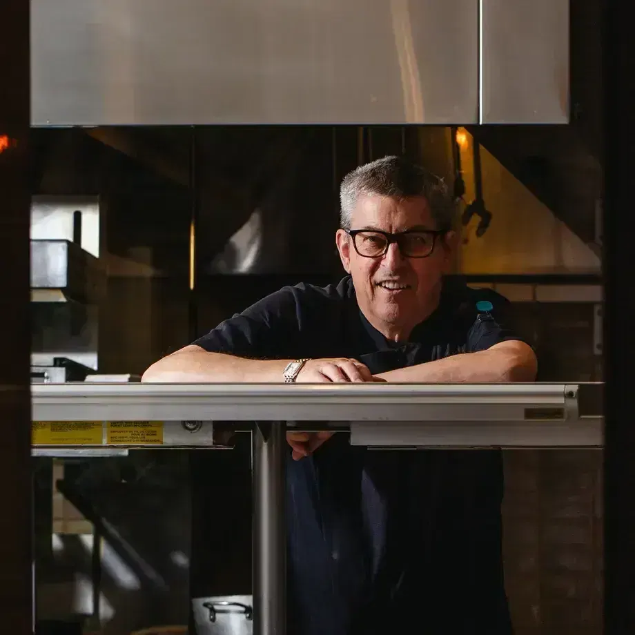 Chef Ken Frank, a middle-aged man with short grey hair and black-framed glasses, smiles while leaning on a stainless steel pass in a professional
