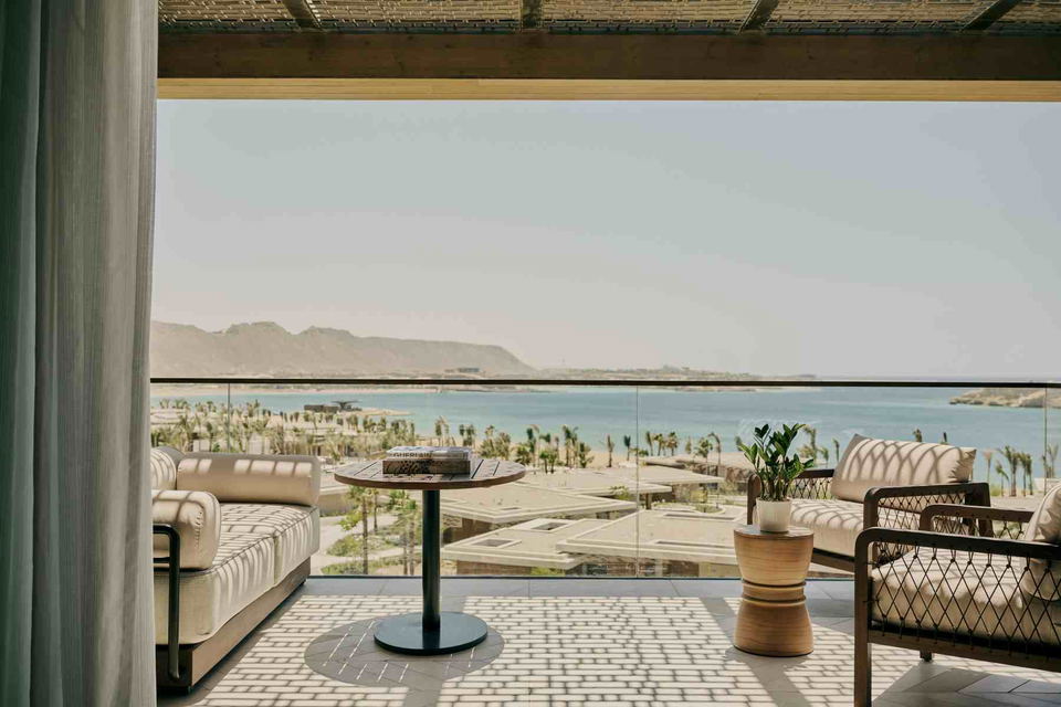 Luxury suite balcony with cream outdoor sofa, round pedestal table with coffee table books, woven side table with potted plant, and glass railing overlooking a palm-lined beach and turquoise Red Sea.