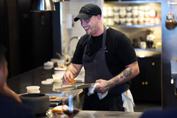 Norman Fenton, a chef wearing a black cap and apron, smiles while holding a small, flat pastry with tongs over a metal tray.