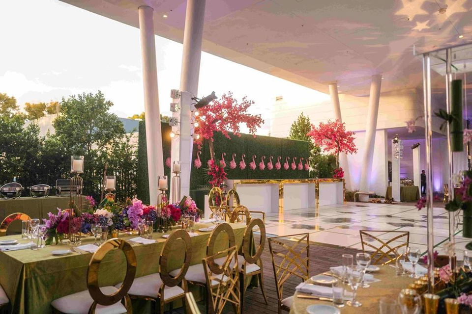 An elegantly decorated outdoor terrace at dusk with gold chairs, green linen tables, vibrant floral centerpieces, a hedge wall with pink flamingo