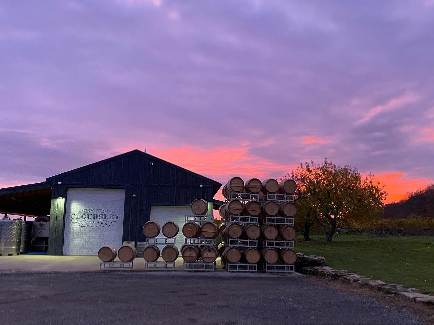 Exterior of Cloudsley Cellars winery building at dusk, with stacked oak wine barrels in the foreground and a vibrant sunset sky.