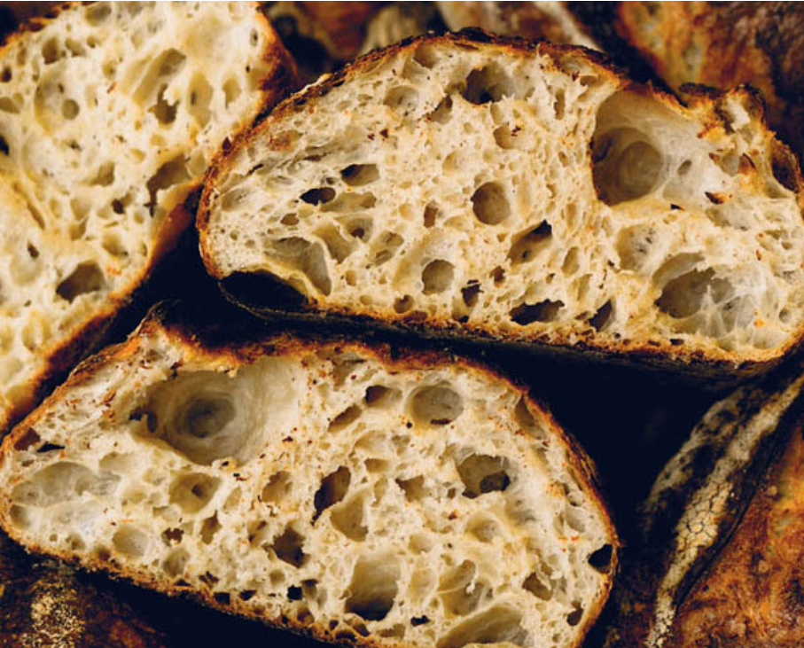 A close-up cross-section of Tartine sourdough bread slices, revealing an open, irregular crumb structure with large air holes and a dark, well-baked
