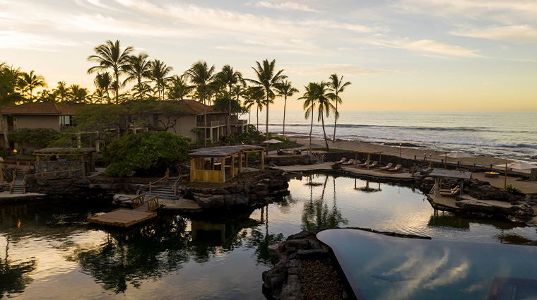An aerial view of the Four Seasons Resort Hualalai at sunset, showing buildings, palm trees, tidal pools, and the Pacific Ocean.