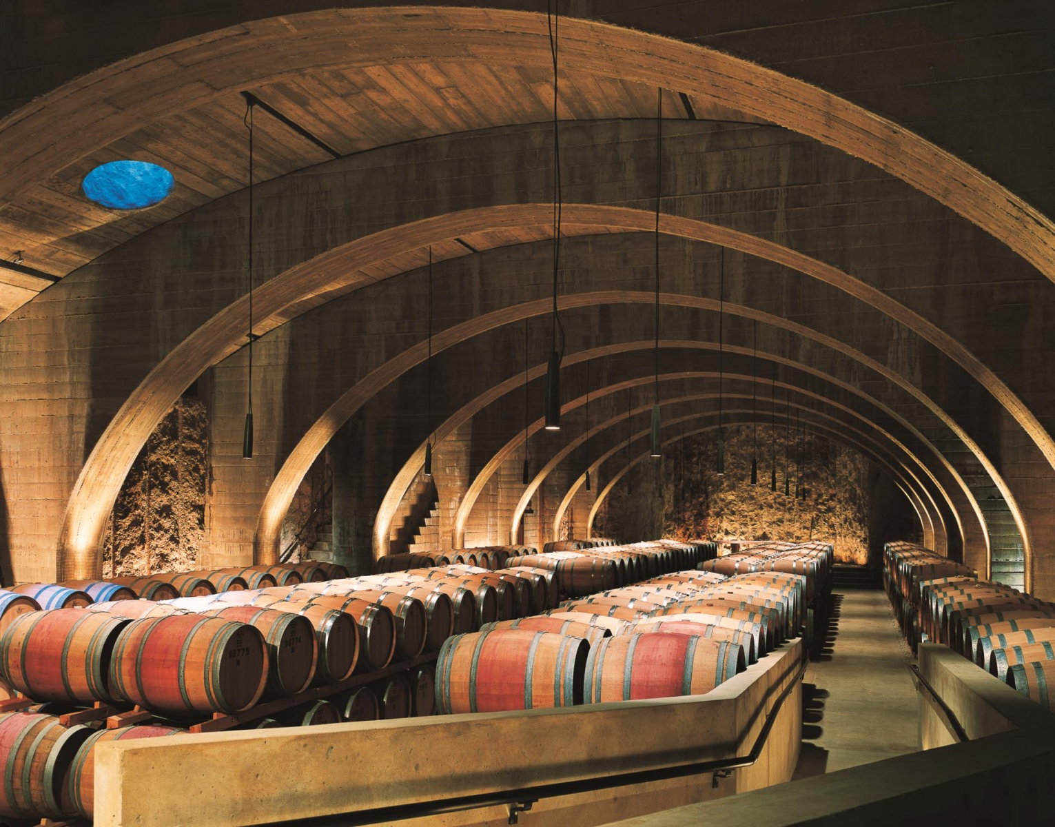 A dramatic barrel cellar with repeated concrete arches, rows of oak wine barrels stacked on both sides, pendant lighting, and a blue oculus skylight.