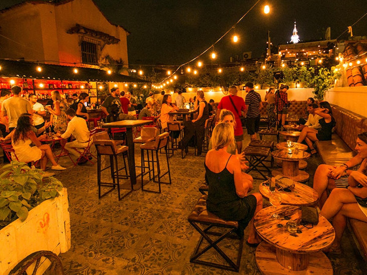 A crowded rooftop bar/terrace at night with string lights, colonial architecture visible in background, and patrons seated at tables; source page expl
