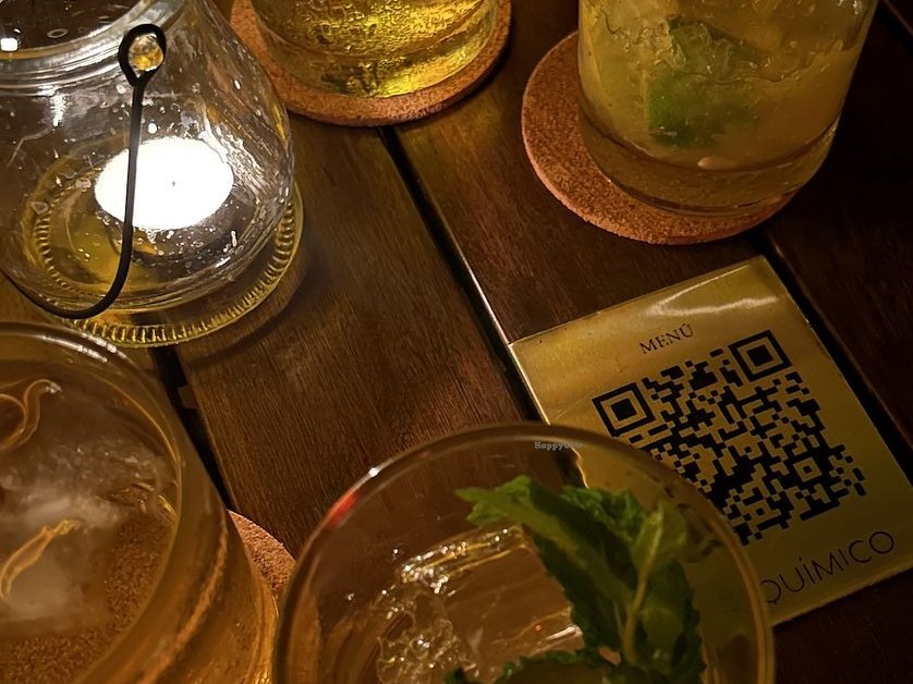 Overhead view of several cocktail glasses with drinks and ice, cork coasters, a lit candle in a glass holder, and a menu card with QR code on a wooden slatted table.