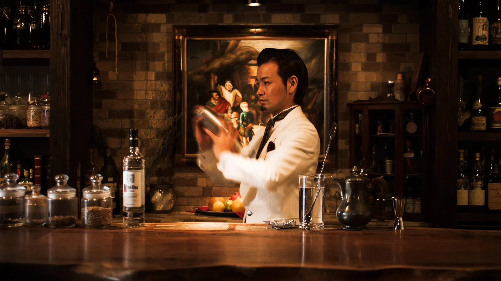 A bartender in a white jacket shakes a cocktail shaker behind a wooden bar, with bottles and jars visible on shelves and the bar.