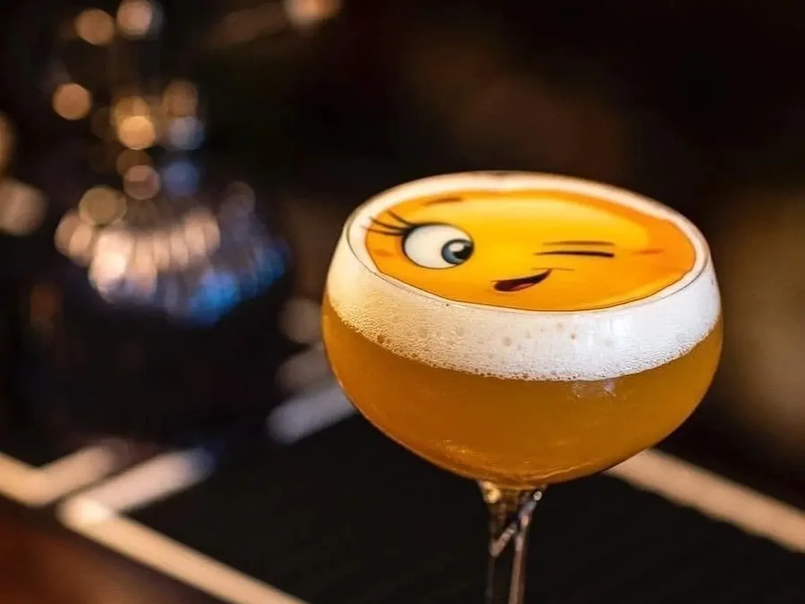 A golden-colored cocktail in a coupe glass with a thick white foam top decorated with a cartoon smiley face design, set against a dark blurred background.