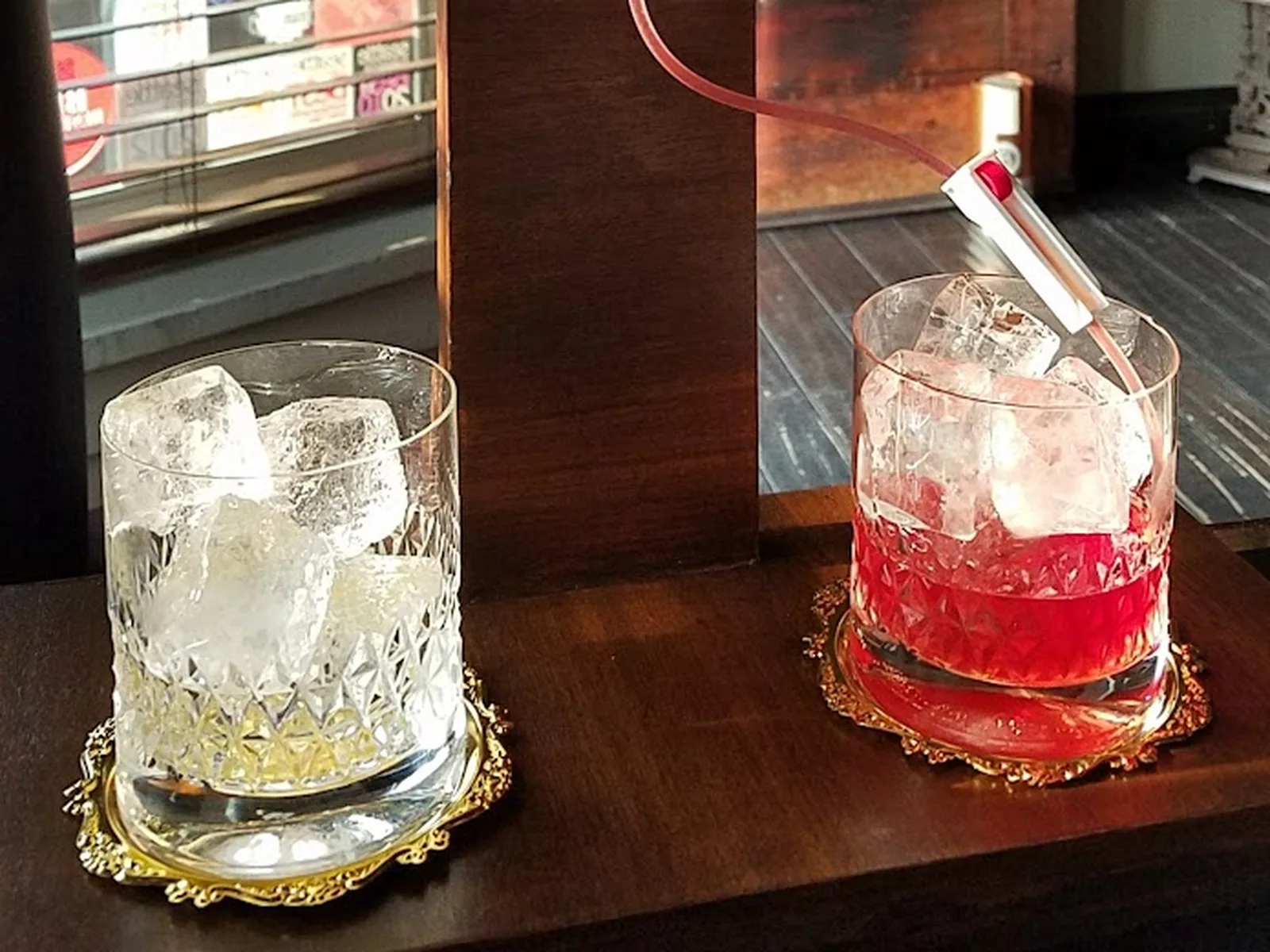 Two cocktails in ornate glasses with ice on brass coasters at a bar, one clear and one red, with a red stirrer in the right glass.