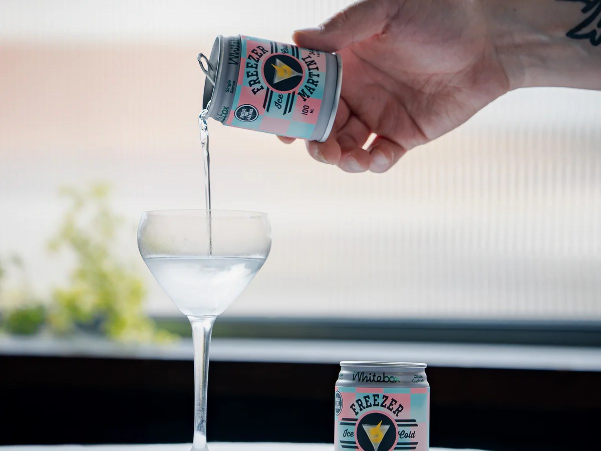 A hand pours a Whitebox Freezer Martini from a small can into a coupe glass, with another unopened can visible on a white table.