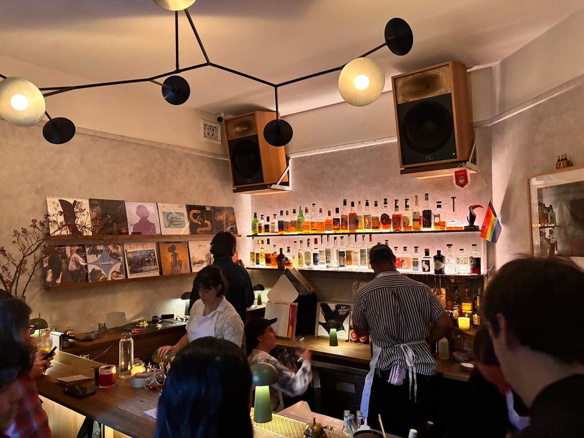 An intimate bar interior with patrons, a bar counter, vinyl records on shelves, backlit liquor bottles, and modern lighting fixtures.