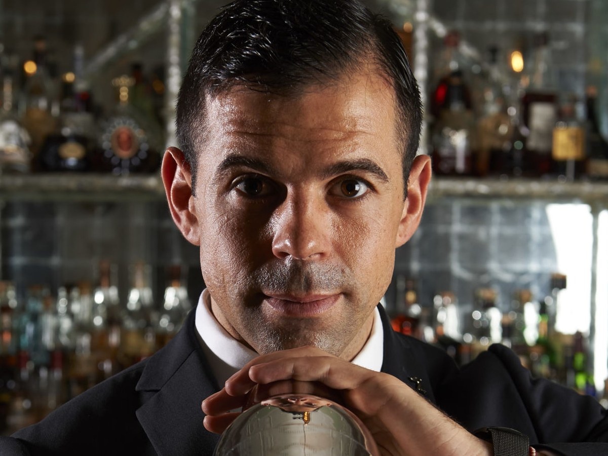 Agostino Perrone, a man with dark hair and a suit, holds a silver cocktail shaker with both hands, looking directly at the camera behind a