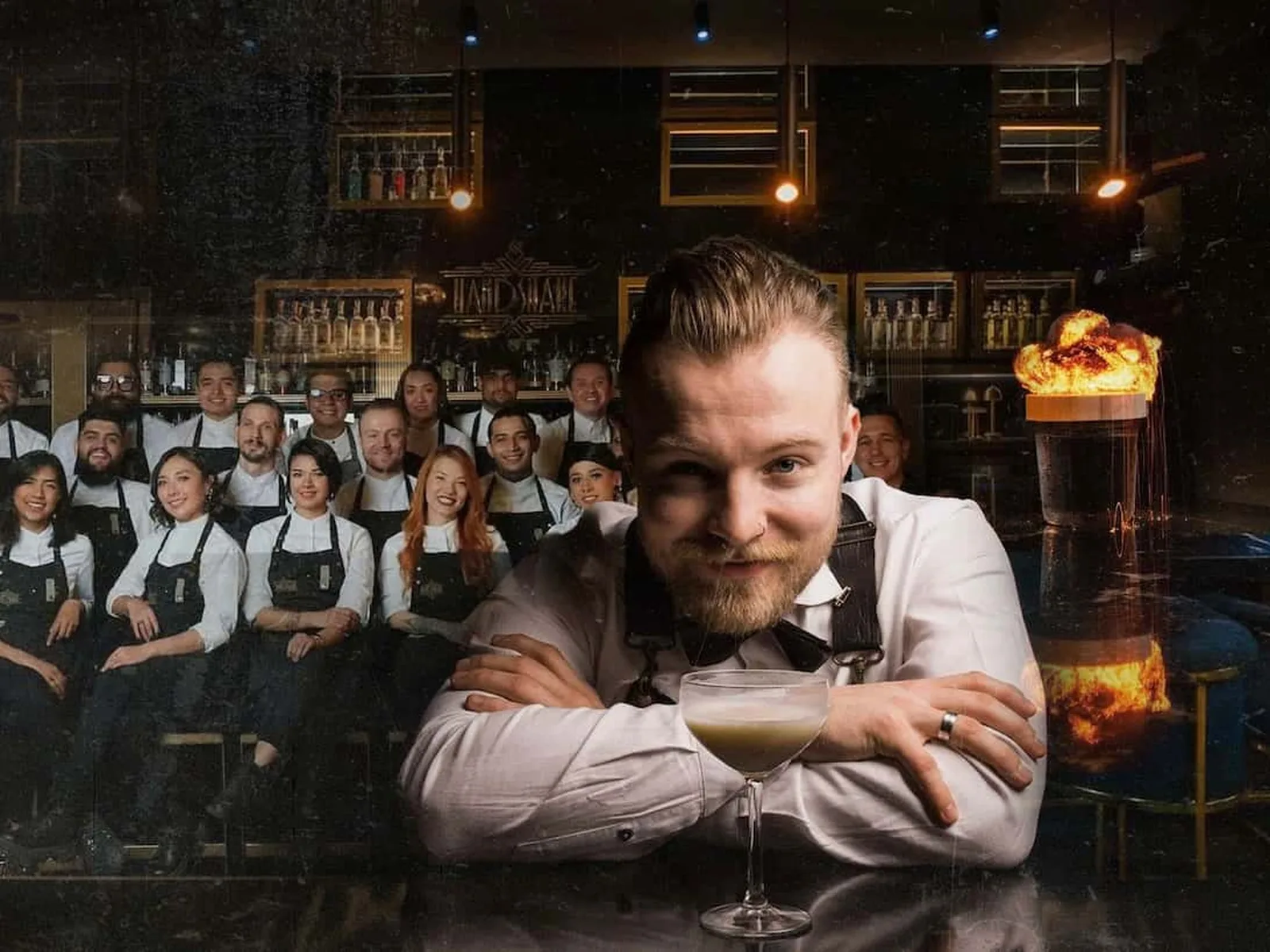 A bartender with a beard and nose ring smiles at the camera, leaning on a bar with a cocktail. Behind him, a group of staff in black aprons pose, and
