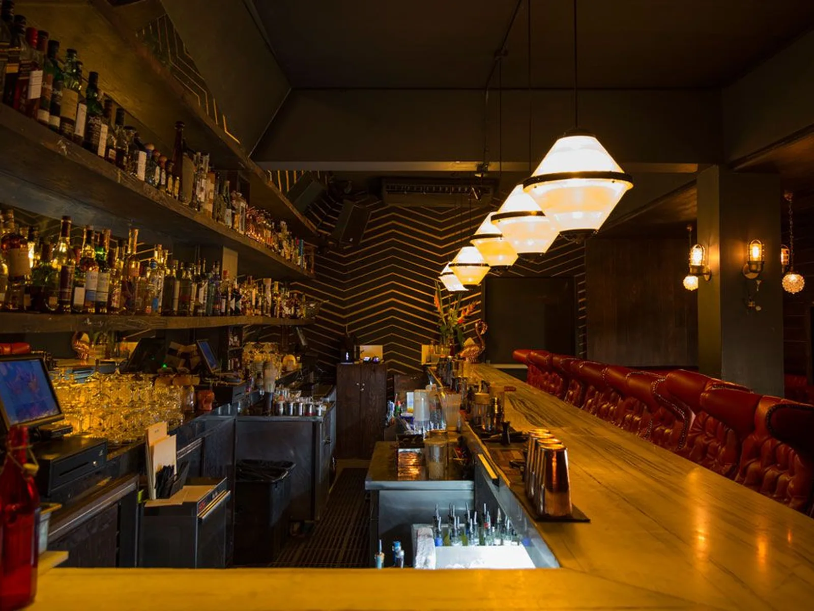 A dimly lit bar interior with shelves of liquor bottles, a long marble bar, and red leather seating. Overhead, several geometric pendant lights