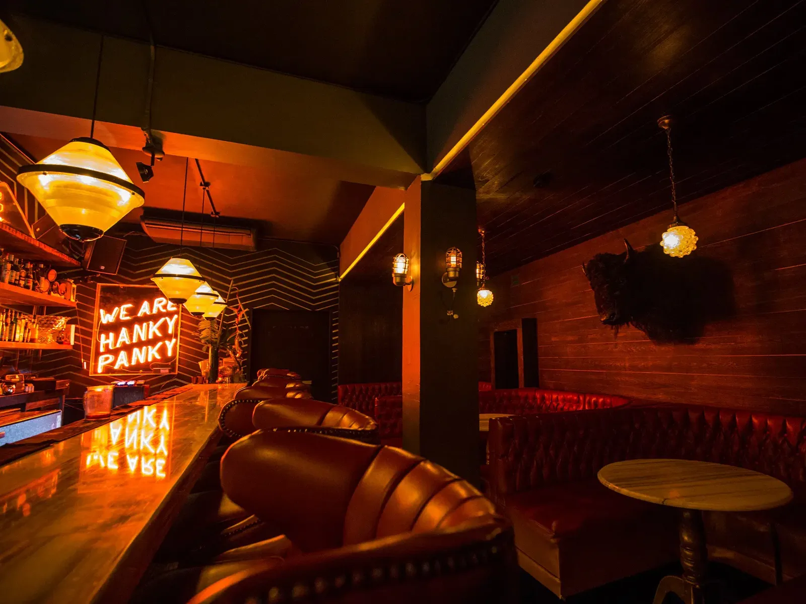 Interior of a dimly lit bar with a long, reflective counter, red leather bar stools, and matching booth seating. A neon sign on the back wall reads