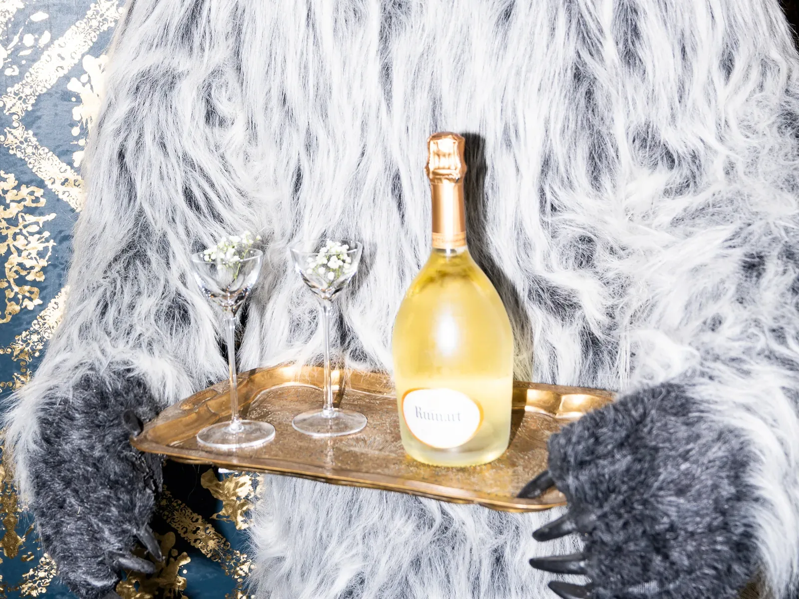 A person in a white and gray furry costume holds a gold tray with a bottle of Ruinart champagne and two glasses filled with baby's breath flowers.