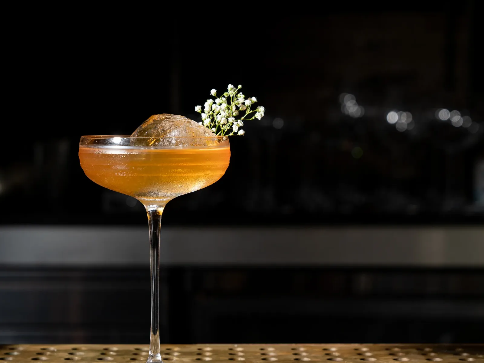 A cocktail in a coupe glass with a large spherical ice cube and a sprig of baby's breath flowers, set against a dark, blurred background.