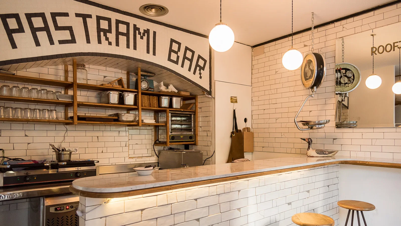 An interior shot of a bar with white tiled walls, wooden shelves, and a marble counter. The words "PASTRAMI BAR" are written on an arched sign above