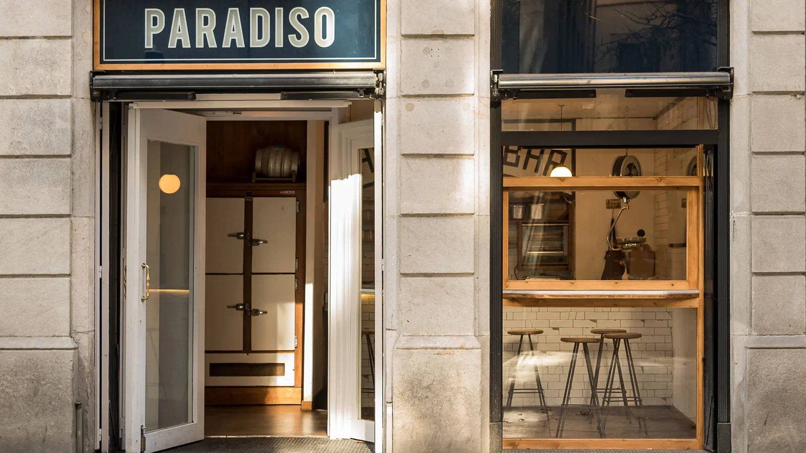 The entrance of Paradiso, a cocktail bar, with a sign above the door and a view into the interior showing a fridge and stools.