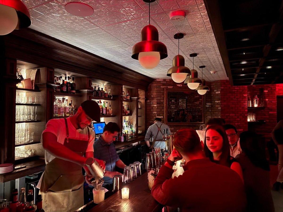 A bartender in a cap and apron pours a drink at a bar with exposed brick walls, a tin ceiling, and red pendant lights.