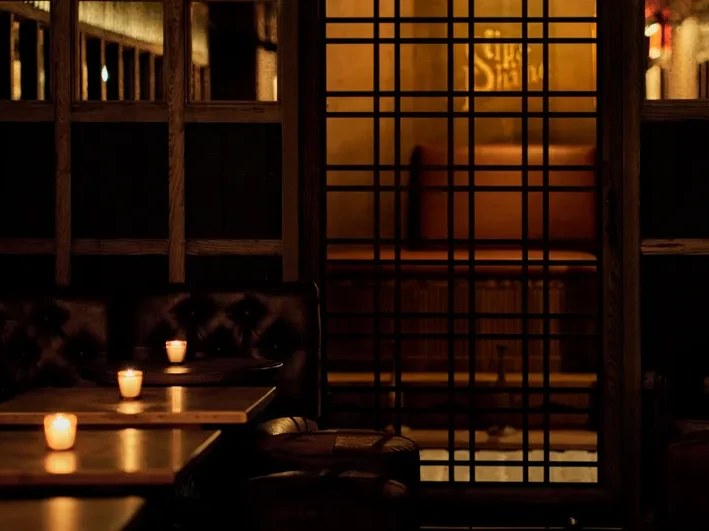 A dimly lit Japanese-style lounge interior with wooden lattice screens, low tables, and glowing candles, creating a moody atmosphere.