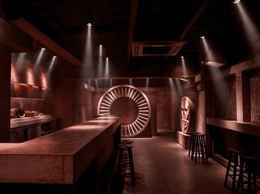 Interior of a bar with dramatic reddish-brown lighting featuring a large circular sculptural disc with radiating elements as the centerpiece, matching