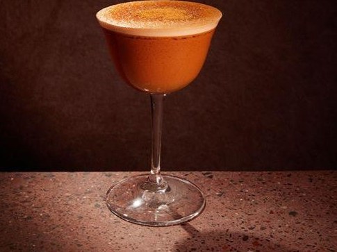 A professionally photographed cocktail in a coupe glass with an orange-brown liquid and foam top dusted with spice, presented on a dark textured surfa