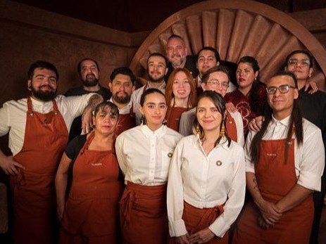 Group portrait of approximately 15 bar staff members in matching rust-colored aprons and white/black shirts, posed in front of a distinctive circular