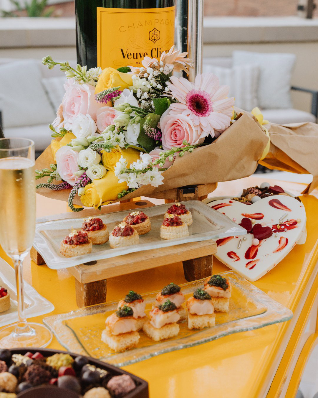Veuve Clicquot champagne, assorted appetizers, floral arrangement, chocolate box on yellow table.