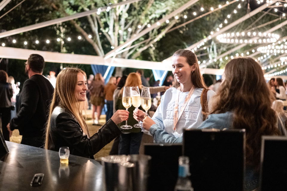 Three guests toast with raised glasses outdoors beneath string lights at a previous Oyster Roast