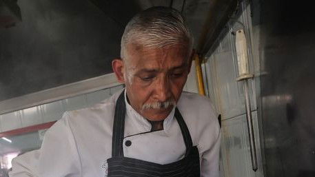 Two cooks prepare food at a busy taquería counter. One cook uses a spatula on a griddle with tortillas and meat, while another operates a tortilla
