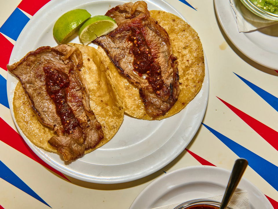 Two steak tacos with lime wedges and red sauce sit on a white plate, against a red, white, and blue striped background.