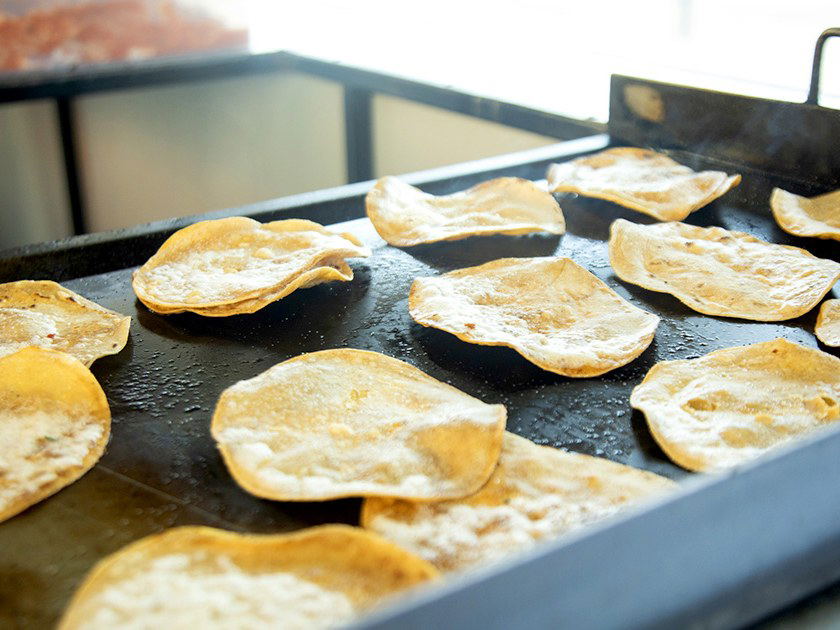 Small, round corn tortillas cook on a large, black flat-top griddle, some lightly browned and puffed up, with visible steam rising.