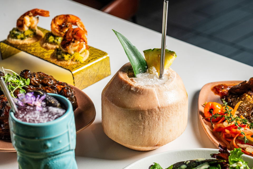 An editorial food and beverage spread showing multiple dishes including shrimp on yellow rice, a coconut drink with pineapple garnish, glazed meat wit
