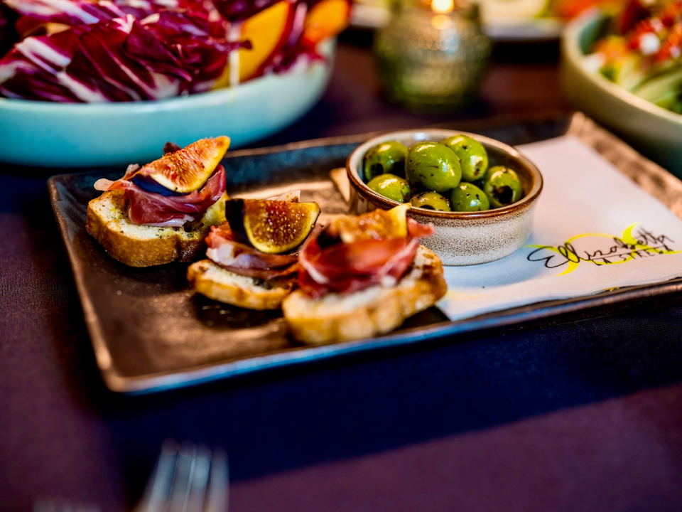 An elegant Italian appetizer platter featuring crostini with prosciutto and figs, alongside marinated green olives in a ceramic bowl, presented on a w