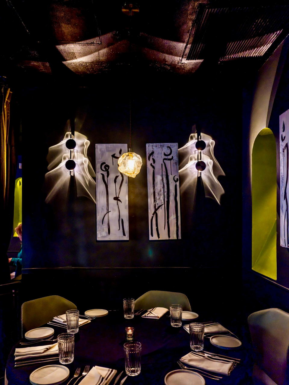 A dimly lit restaurant dining room with two place settings, white sculptural hanging lights on either side, two framed artistic panels with black figure drawings in the center, and a golden glowing pendant light between them.