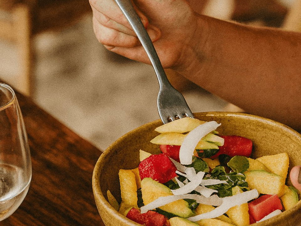 A person holds a fork over a bowl of fresh fruit salad with watermelon, pineapple, cucumber, and shredded coconut.