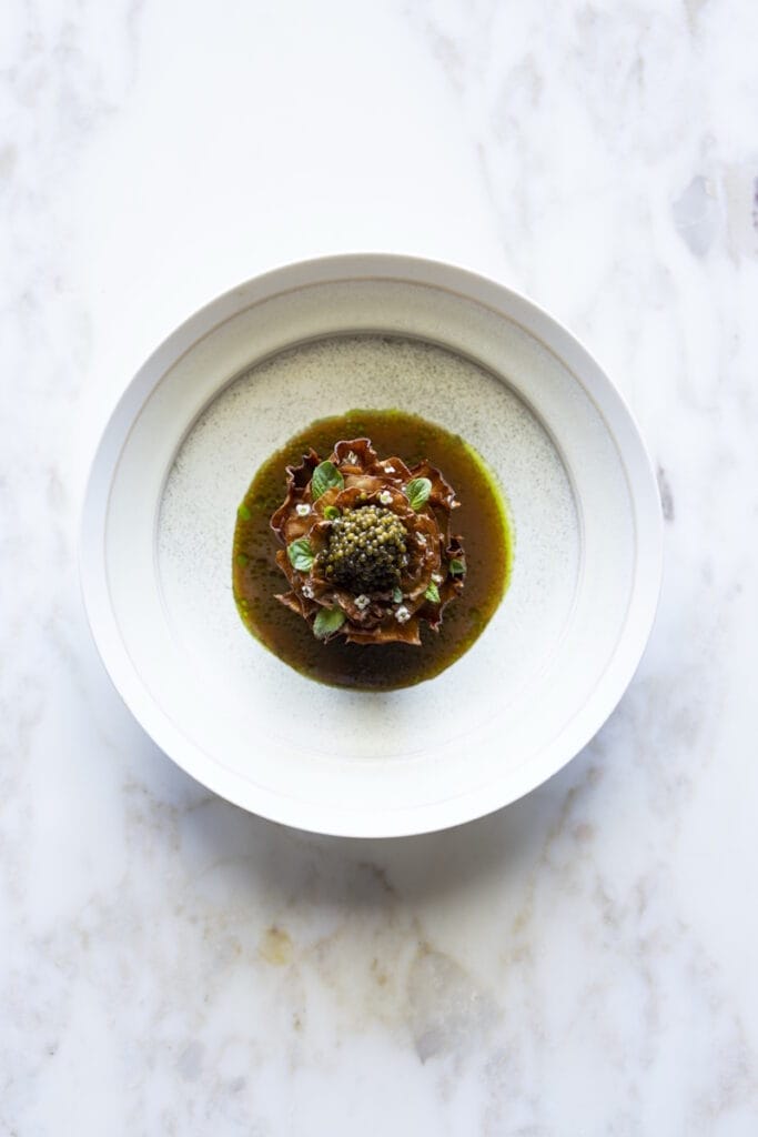 High-end plated dish showing artichoke with caviar on elegant ceramic, presented with decorative greenery wreath; source page from Elite Traveler expl