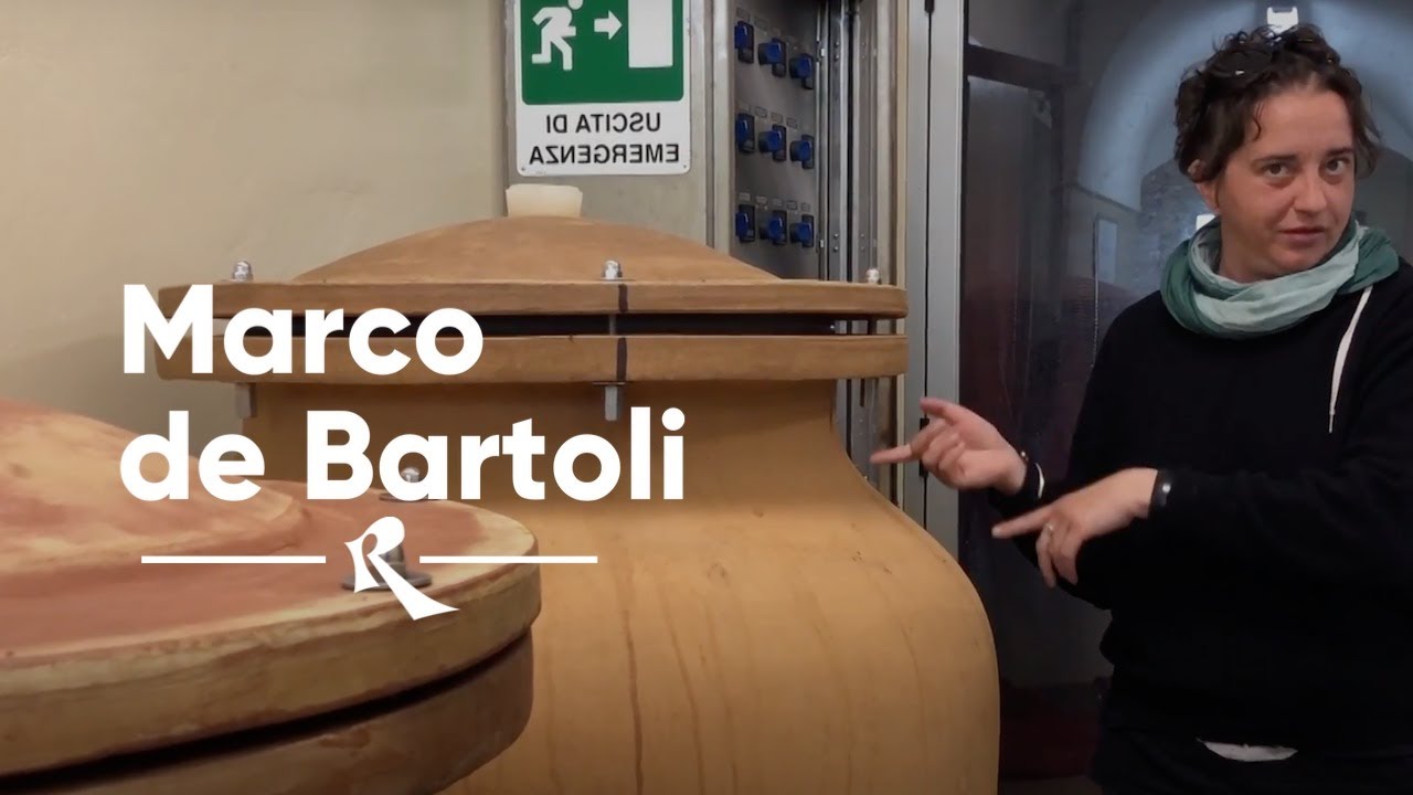 A person gesturing toward large wooden barrels in a cellar, with visible text overlay reading 'Marco de Bartoli'