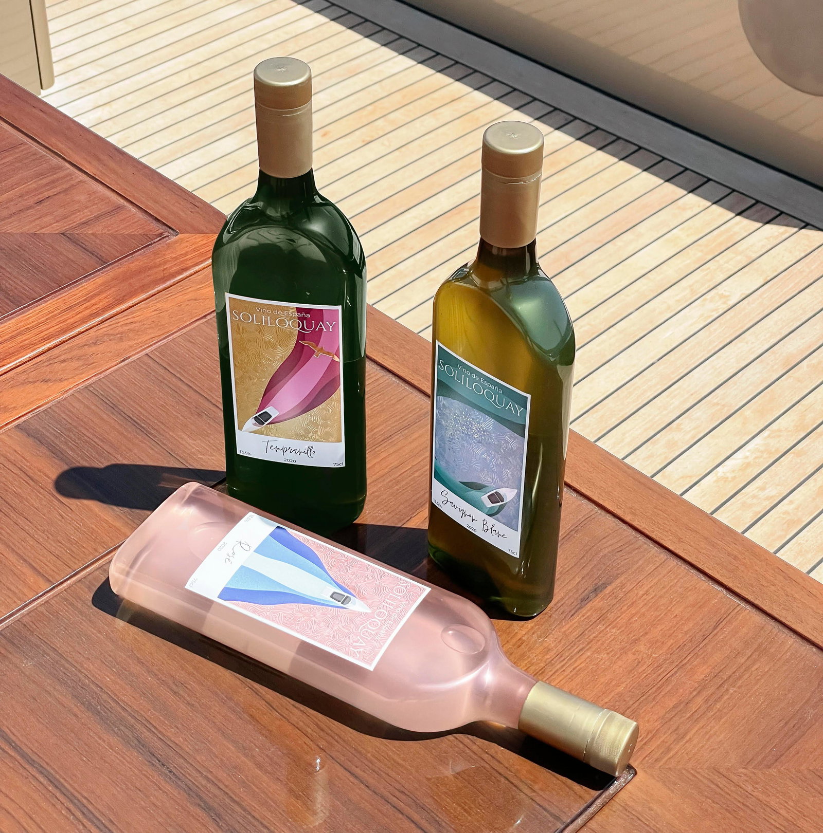Three wine bottles with bold, modern illustrated labels (Soliloquay brand) arranged on a wooden deck surface.