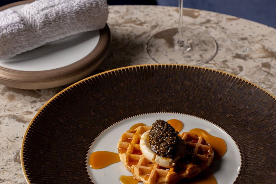 A waffle topped with a dollop of white cream and a generous serving of black caviar, drizzled with a golden sauce on a decorative plate.